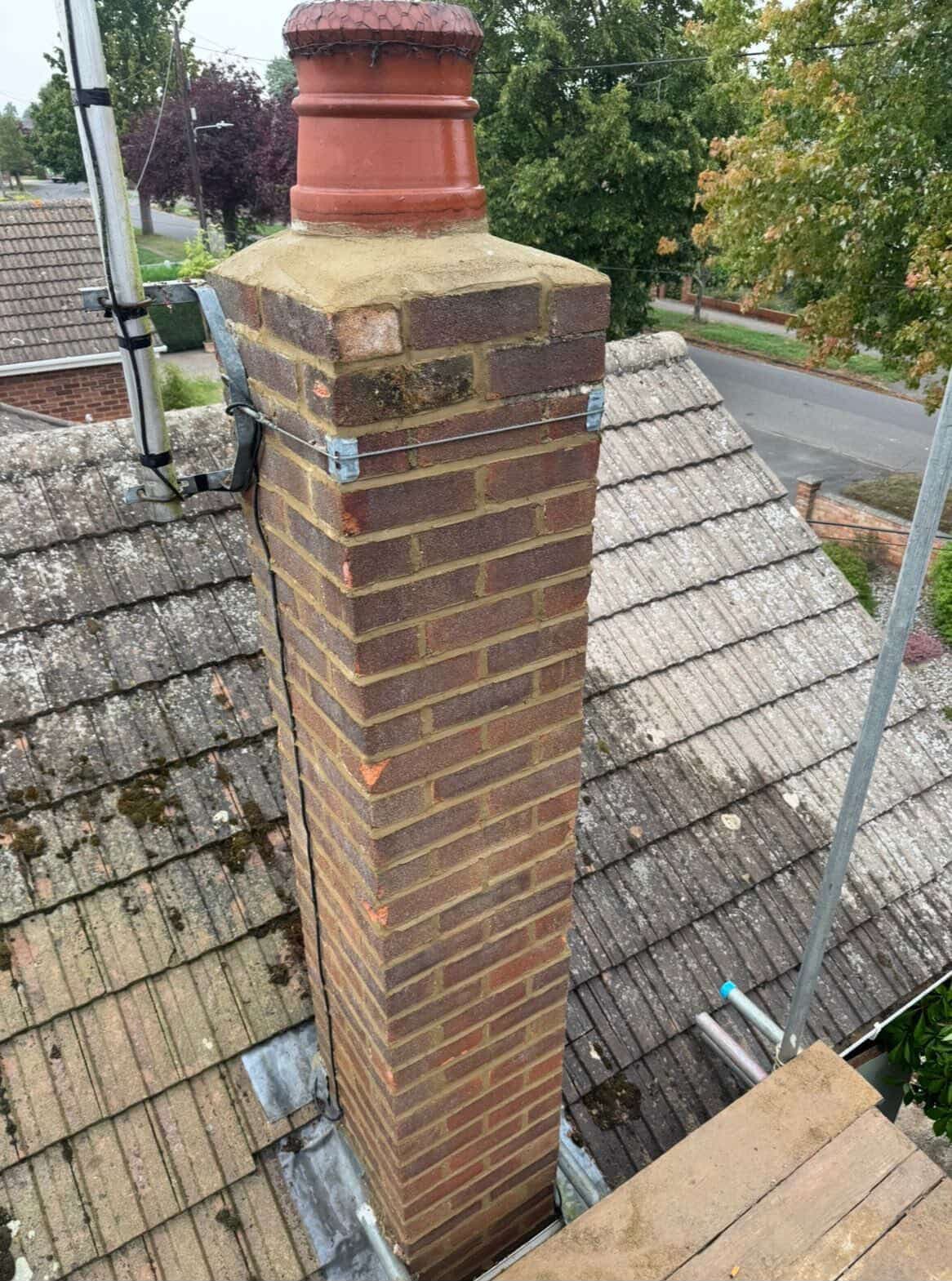 Tall brick chimney with a clay pot and antenna on a weathered tiled roof.