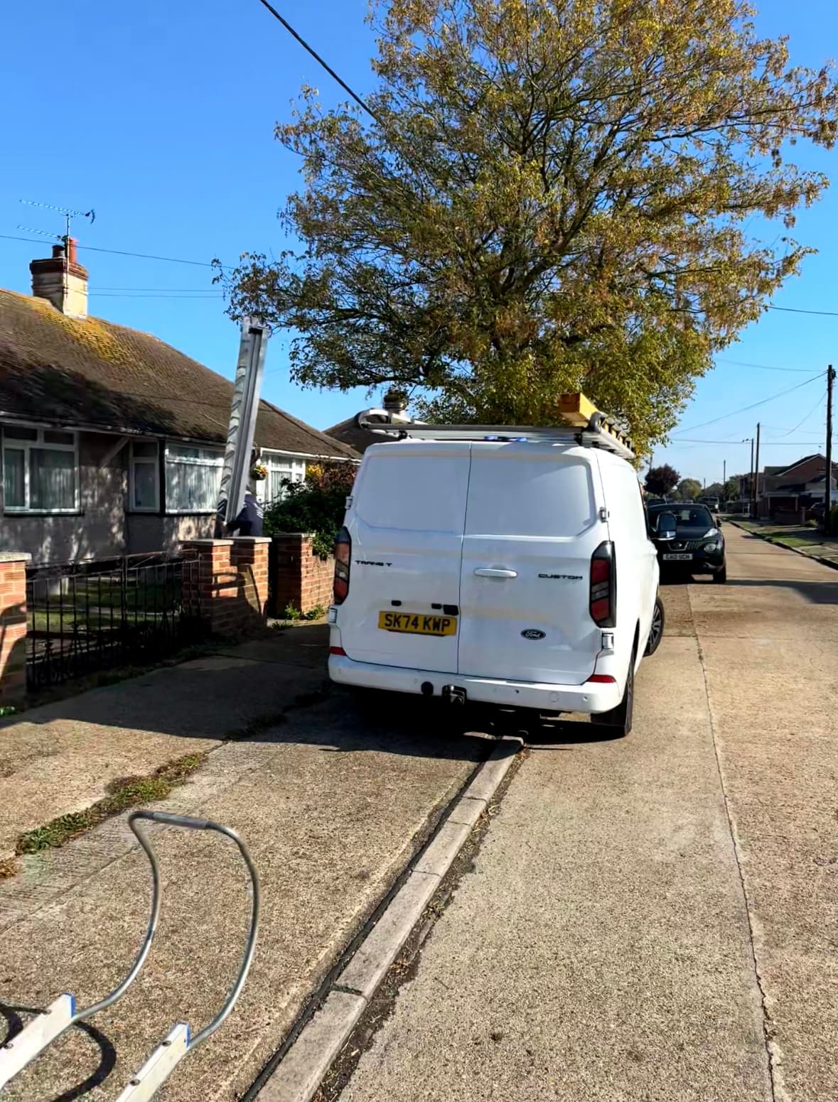 White Ford Transit van with roof ladders parked on a sunny residential street.